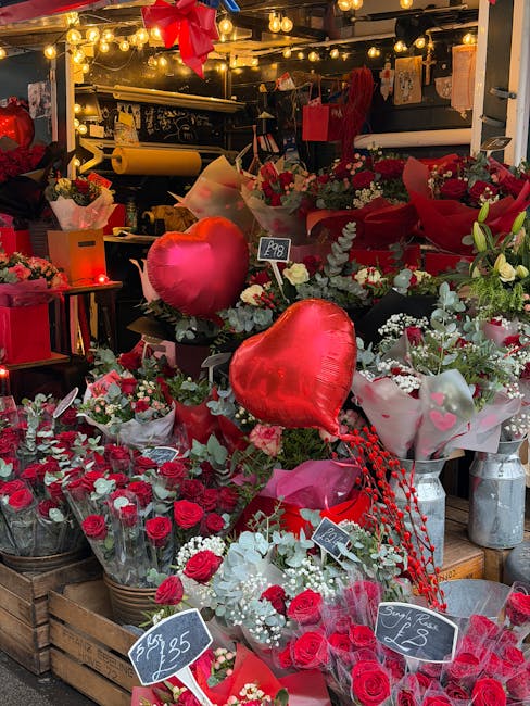 An indoor floral display featuring an assortment of fresh roses, predominantly in red and white, arranged in bouquets and decorative wrapping. The arrangement includes heart-shaped helium balloons in 