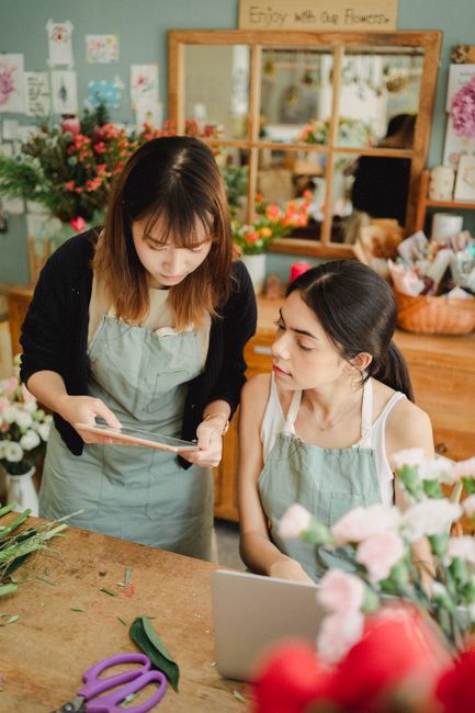 Inside a flower workshop at East Dulwich florist, two women are engaged in a floral arrangement task. The woman on the left, with shoulder-length brown hair and wearing a black cardigan over a light-c