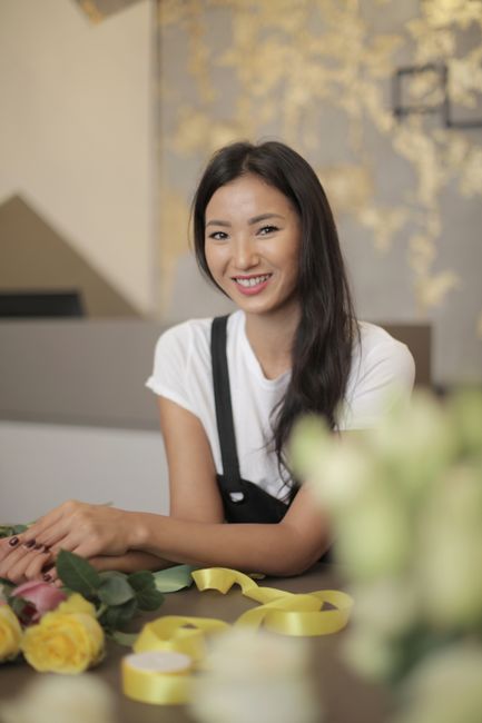 A smiling young woman with long dark hair wearing a white t-shirt and black apron, sitting at a reception desk with a blurred background of floral artwork on the wall. In front of her are fresh flower