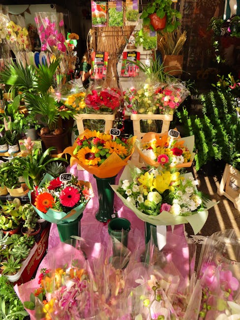 A vibrant display of fresh floral bouquets arranged on a pink table within a flower shop. The bouquets feature a variety of colorful flowers including bright orange gerbera daisies, yellow sunflowers,