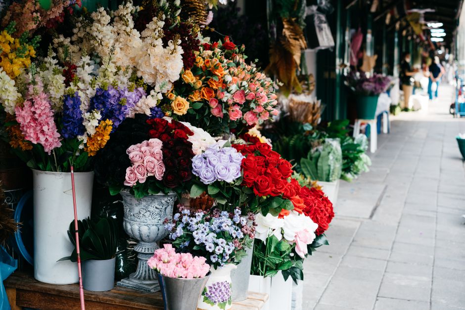 A vibrant display of fresh flowers arranged in various vases along the sidewalk of a flower shop, featuring roses in shades of pink, red, and white, as well as iris and stock blooms in purple, white, 
